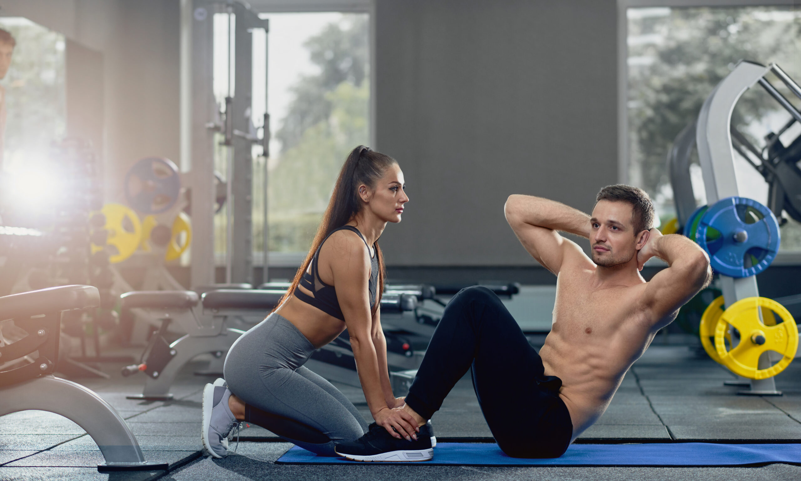 Side view of sexy woman wearing in sportswear, holding man feet down as he doing sit up exercises. Man doing abdominal crunches press exercise on mat with bare torso in gym. Teamwork concept.